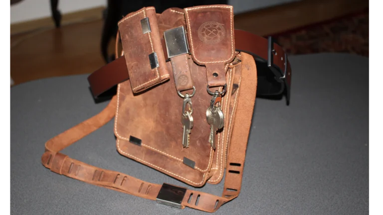 Fotografie care prezintă familia de produse dezvoltată de Ludor Engineering împreună cu clientul și partenerii. Imaginea surprinde un ansamblu de accesorii din piele maro, așezate pe o suprafață gri texturată. În centru se află o geantă de tip buzunar sau toc, prevăzută cu două clapete distincte și un sistem inovator de prindere din metal. De geantă sunt atașate, prin intermediul unor inele metalice, două brelocuri cu chei. În prim-plan se află o centură din piele, cu un mecanism metalic de blocare rapidă și mai multe fante dreptunghiulare pentru ajustare. Pe fundal este vizibilă o a doua centură de culoare mai închisă, cu catarame metalice. Toate piesele sunt realizate din piele naturală, cu cusături albe vizibile, și integrează elegant componente metalice, demonstrând o linie de produse coerentă, funcțională și gata pentru utilizare.