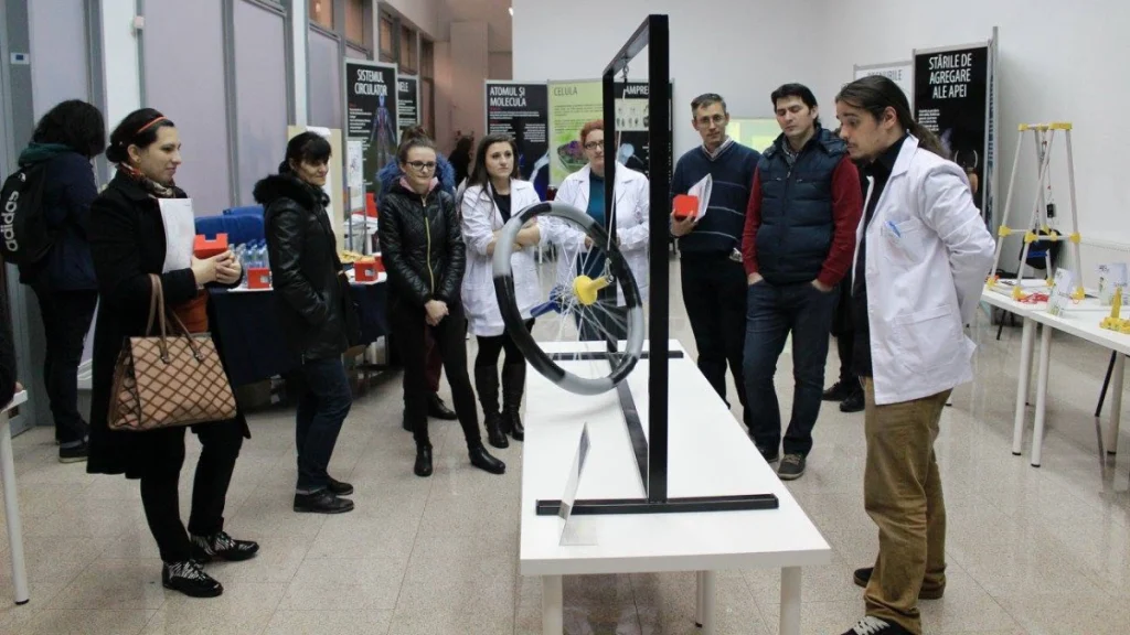 Fotografie care surprinde lansarea expoziției de știință interactive Laborator, desfășurată într-o sală spațioasă, tip muzeu, cu pereți albi și panouri informative pe teme științifice (vizibile în fundal: „SISTEMUL CIRCULATOR”, „ATOMUL ȘI MOLECULA”, „STĂRILE DE AGREGARE ALE APEI”). În centru, pe o masă albă, se află un exponat fizic proiectat și fabricat de Ludor Engineering: o roată de bicicletă montată vertical într-un cadru de susținere, cu un ax și un element galben, care demonstrează principii de fizică (precum precesiunea giroscopică sau conservarea momentului cinetic). În jurul mesei, un grup numeros de vizitatori (adulți și tineri) privesc cu interes exponatul. Câțiva dintre ei poartă halate albe de laborator, având rolul de ghizi sau facilitatori. Un tânăr din dreapta, îmbrăcat în halat alb, pare să explice participanților mecanismul de funcționare al instalației.