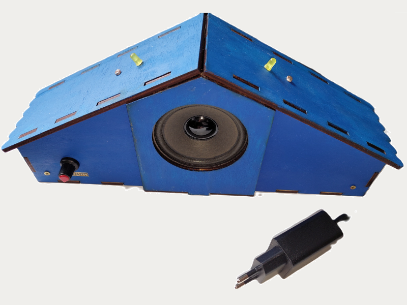 Fotografie a unui theremin educațional (instrument muzical electronic) dezvoltat și fabricat de Ludor Engineering, pe un fundal alb. Dispozitivul are o formă de căsuță, fiind construit din placaj de lemn vopsit în albastru, cu îmbinări prin tăiere. În centrul feței frontale este montat un difuzor negru. Pe panoul din stânga se află două butoane rotative (unul negru și unul roșu) și o mică plăcuță aurie cu denumirea butonului. Pe suprafețele superioare înclinate se observă doi senzori LDR (rezistențe dependente de lumină) și 2 LED-uri galbene. Senzorii controlează sunetul prin umbra mâinii. Alături, în partea dreaptă jos, se află un adaptor de priză (încărcător) negru folosit pentru alimentarea dispozitivului. Imaginea ilustrează un produs inteligent care integrează senzori, componente electronice (Arduino) și design mecanic, potrivit pentru educație.