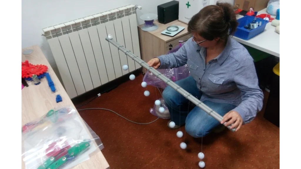Fotografie care ilustrează Etapa 4 – Prototip: prototipare rapidă și probe funcționale, desfășurată în atelierul Ludor Engineering. O femeie ține în mâini un prototip de formă alungită, asemănător unui pendul, format dintr-o tijă argintie segmentată. De tijă sunt suspendate mai multe bile albe, atârnând la diferite înălțimi, printr-un sistem de fire. Femeia verifică atent echilibrul și funcționarea mecanismului, purtând ochelari de protecție. În stânga imaginii, pe birou și pe podea, se află diverse piese imprimate 3D (în culori roșu, verde și albastru), unelte, o rolă de plastic transparent și componente metalice. Imaginea surprinde procesul de asamblare de probă și primele verificări mecanice și ergonomice.